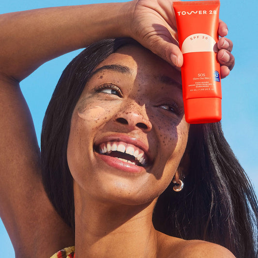 A person smiles while holding up a bottle of Tower 28 Beauty's SOS FaceGuard SPF 30. A swatch of the product is visible on their cheek. [Shared: SOS FaceGuard SPF 30]